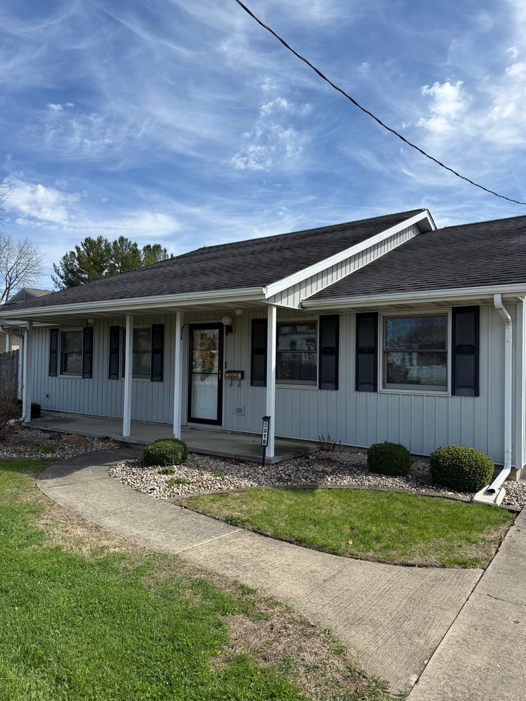White ranch-style home with front porch, dark roof, concrete driveway, and landscaping on a sunny day