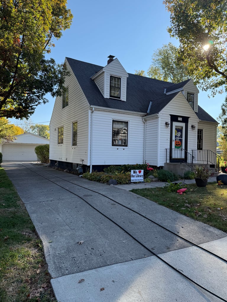 White Cape Cod style house with black roof and dormers, front porch, driveway with railroad tracks, surrounded by trees