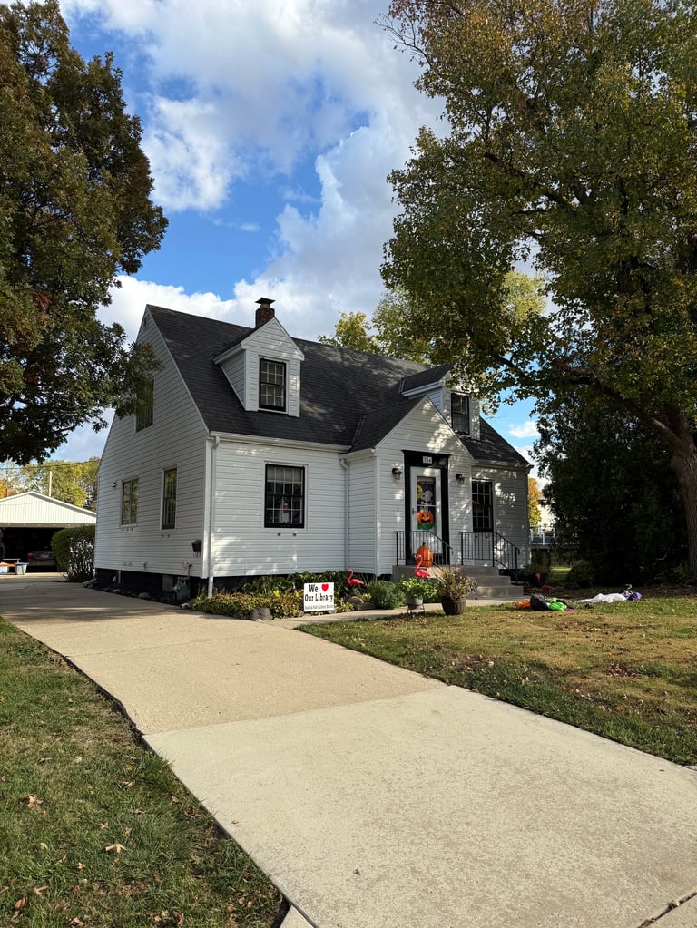 White cottage-style house with black roof and front porch surrounded by tall trees and green lawn