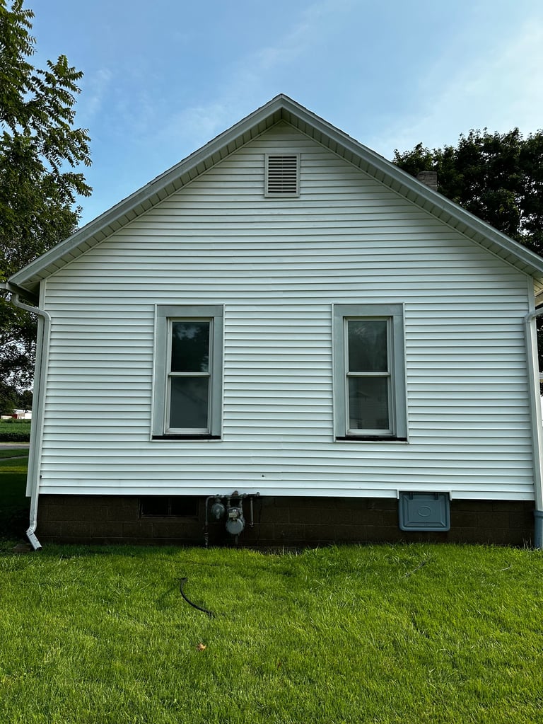 Small white single-story house with horizontal siding, two front windows, and gabled roof surrounded by green lawn and trees