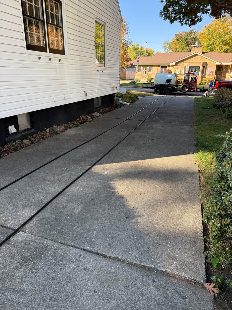 Driveway with black electrical lines running along a white mobile home, with yard and neighboring buildings in background