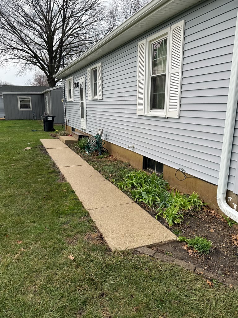 Single-wide mobile home with light blue siding, concrete walkway, landscaping beds, and bare tree in yard