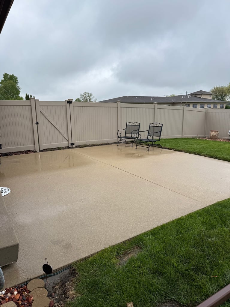 Backyard patio with concrete floor, two lounge chairs, metal fence, and surrounding grass under cloudy sky