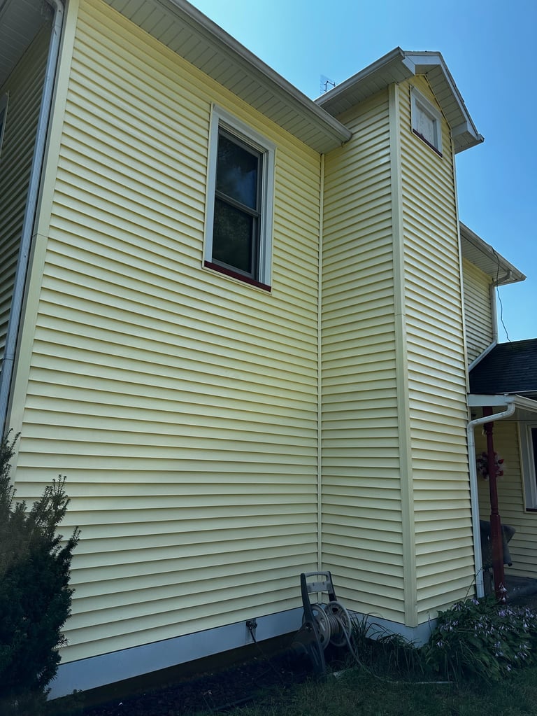 Two-story yellow house with horizontal siding, dark window, and peaked roofline against clear blue sky