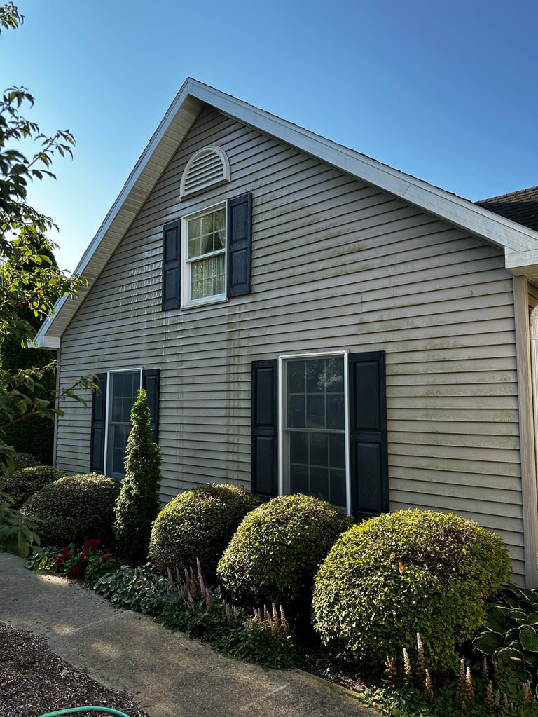 Gray house with gable roof, dark shutters, and round topiary bushes lining the front foundation against clear blue sky