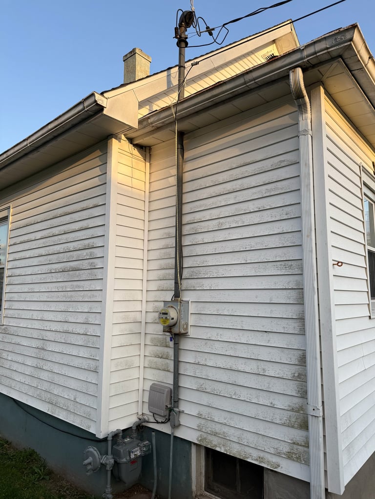 Exterior of a small white and gray house with horizontal siding, electrical meter, power lines, and roof overhang visible