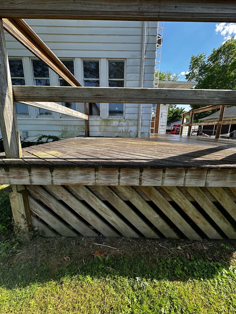 Wooden deck under construction attached to white house with beams and railings, surrounded by green landscaping and trees