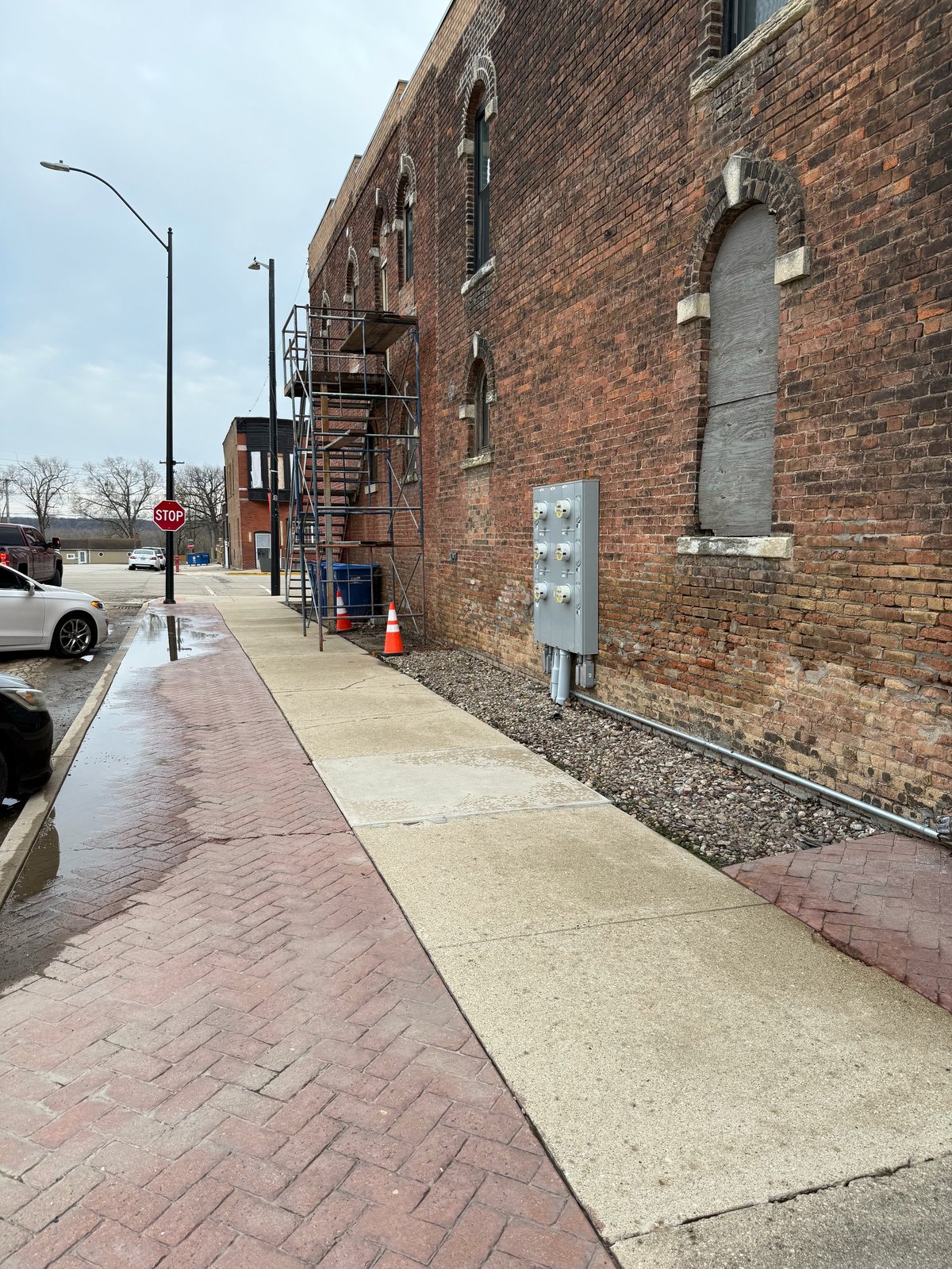 Urban brick building exterior with adjacent sidewalk, fire escape, street lamp, and parked car visible on a partly cloudy day