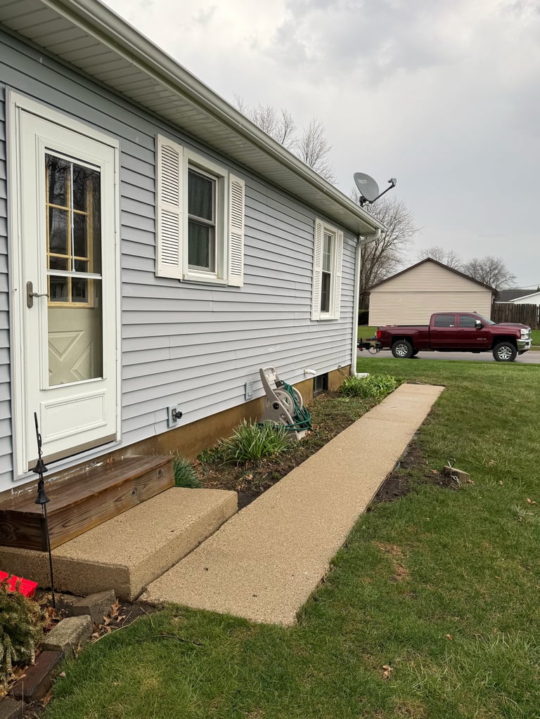 Single-wide mobile home with white vinyl siding, concrete walkway leading to front entrance, and parked maroon pickup truck in background