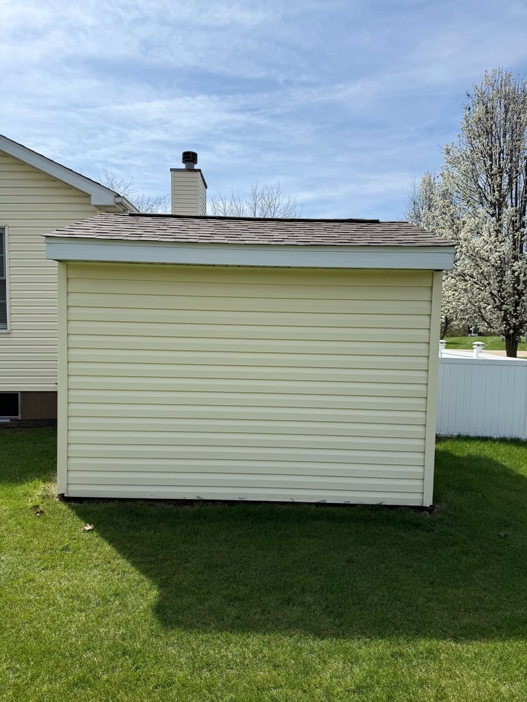 Beige vinyl-sided detached garage or storage shed on green lawn with chimney and metal roof
