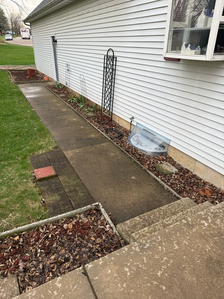 A concrete walkway alongside a white-sided house with landscaped mulch beds, black trellis, and green lawn