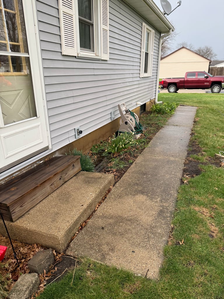 Concrete walkway along side of house with gray siding, landscaping beds, and red pickup truck visible in background