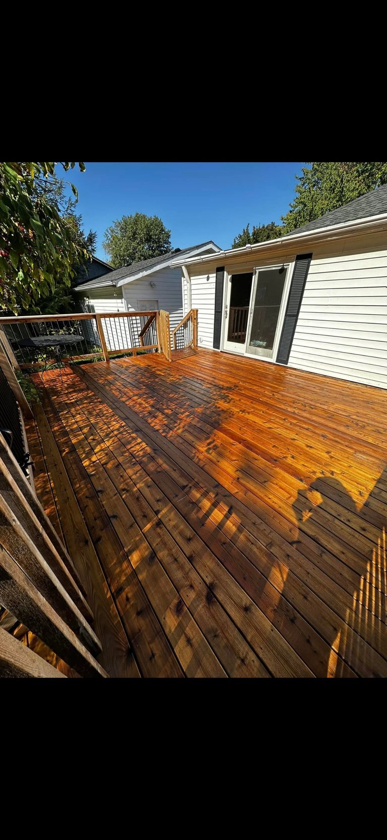 Large wooden deck attached to white house with railing, in sunny backyard with blue sky and green trees