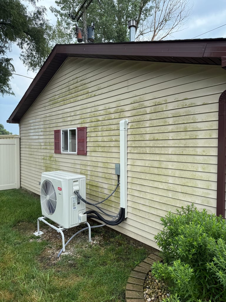 White air conditioning unit mounted on the exterior wall of a beige vinyl-sided house with a dark roof and burgundy window shutters